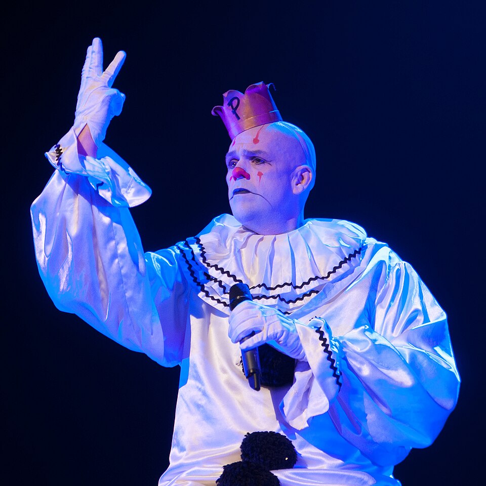 Puddles the Sad Clown, in white satin clown suit and small gold crown, singing into a microphone on a dark stage