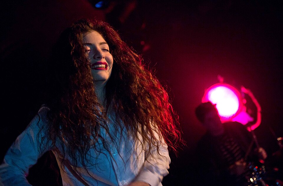 Lorde, with long curly hair and a white shirt, smiling on stage under red lights in 2013