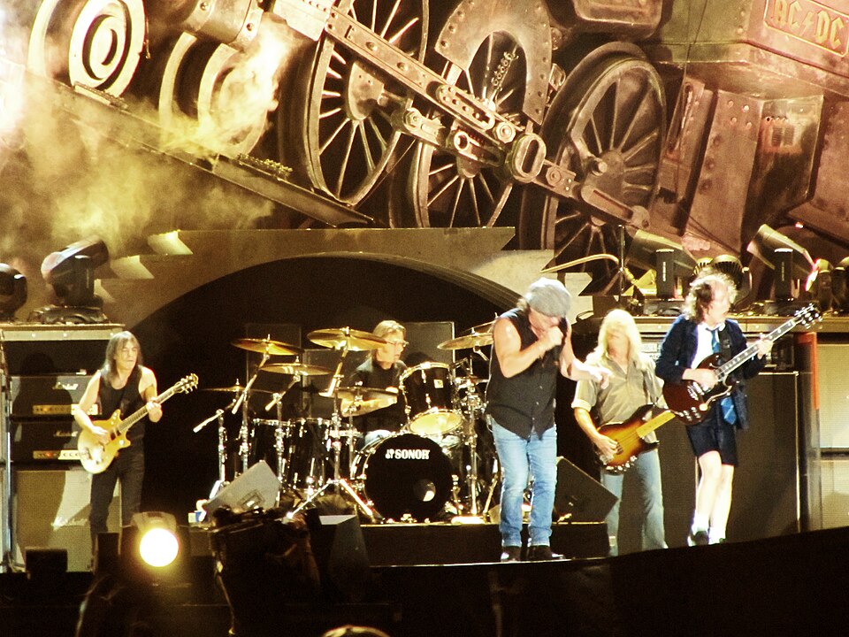 AC/DC on stage in front of a giant locomotive set piece, with Angus Young in his schoolboy uniform and Brian Johnson in a flat cap