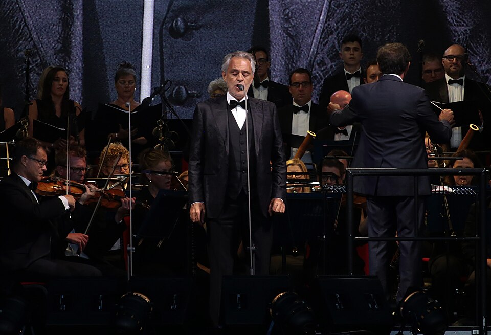 Andrea Bocelli in a black tuxedo singing on stage in front of an orchestra and choir, with the conductor at right