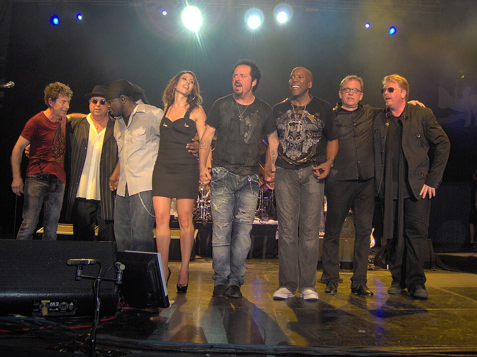 The eight members of Toto standing arm in arm at the front of a concert stage, taking a bow