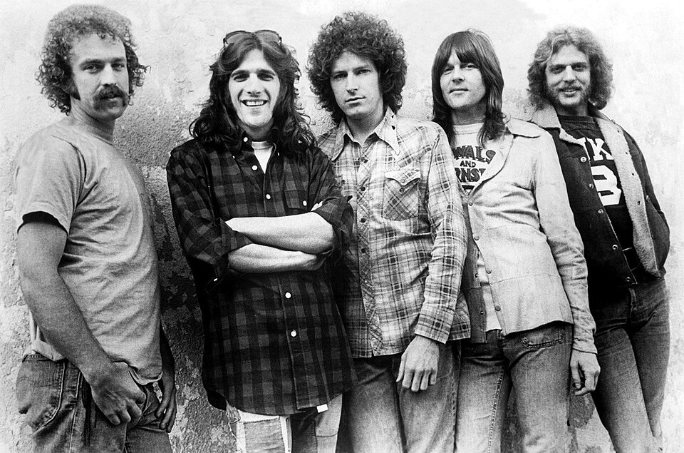 Black-and-white photo of the five members of the Eagles in jeans and flannel shirts, leaning against a stone wall