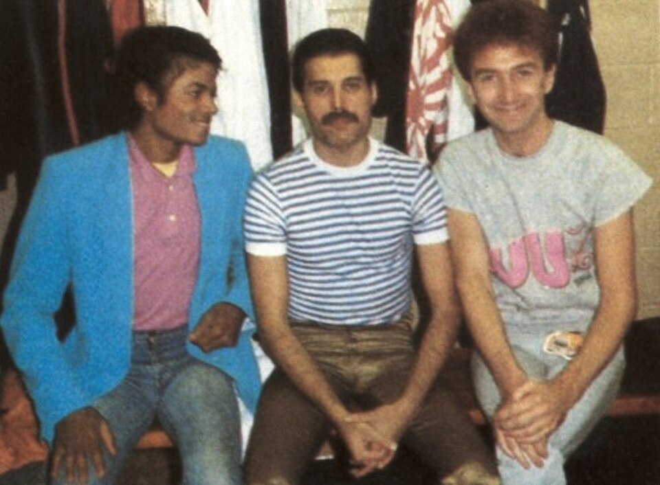 Michael Jackson in a blue blazer seated backstage beside Freddie Mercury in a striped T-shirt and Queen bassist John Deacon, 1982
