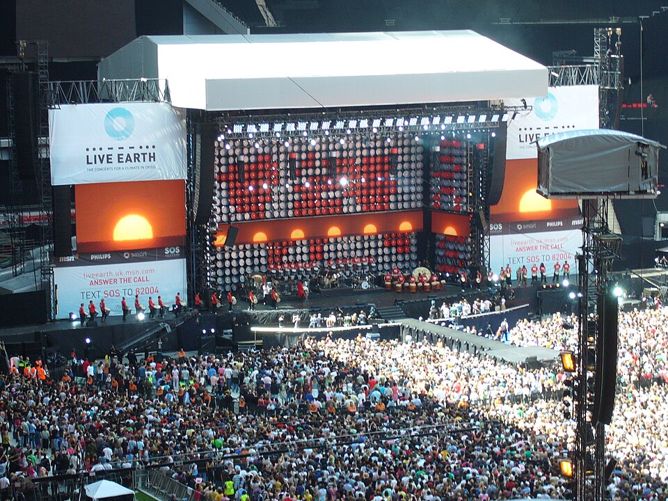 A packed Wembley Stadium crowd facing the Live Earth stage, with orange sun graphics on the screens