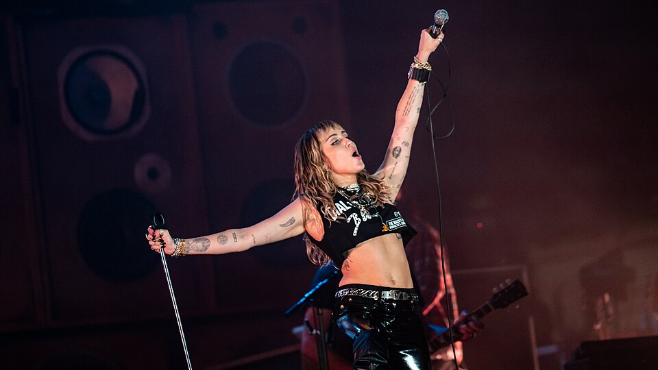 Miley Cyrus on stage with one arm raised holding a microphone, in a black crop top and leather pants