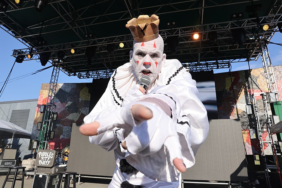 Puddles the sad clown in white clown suit and gold crown reaching a gloved hand toward the camera on an outdoor festival stage